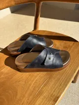 Sandalia de cuero con diseño de tiras cruzadas en color azul oscuro, detalle de doble remache dorado y plataforma de madera con plantilla metalizada.