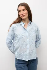 Camisa celeste con apliques florales bordados, cuello camisero, manga larga y cierre frontal de botones.