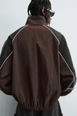 Cazadora de ajuste relajado, color marrón oscuro con bloques de color negro en las mangas. Presenta ribetes blancos que delinean los paneles y cuello alto con cierre frontal de cremallera. Los puños y el bajo son elásticos.