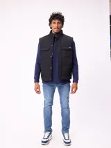 Chaleco acolchado color negro con cierre frontal de cremallera y botones a presión. Presenta cuello alto, bolsillos frontales con solapa y un pequeño parche con logo en el pecho.