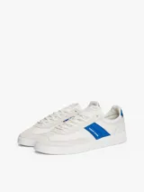 Zapatillas deportivas de estilo vintage Tommy Jeans modelo The Greenwich, confeccionadas en ante color blanco con paneles laterales en azul vibrante y detalles en gamuza gris claro. Presentan cierre con cordones, suela cupsole de goma y logo de la marca en el lateral.