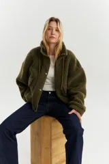 Campera de sherpa color verde musgo, con cuello alto, cierre frontal de cremallera y solapa con botones a presión. Presenta bolsillos delanteros con solapa y cordón de ajuste en el ruedo.