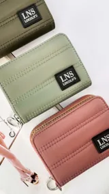 Billetera corta de diseño capitoneado con cierre perimetral y tapa frontal. Cuenta con compartimentos internos para tarjetas y billetes, e incluye una correa de mano desmontable. Presenta un aplique de goma con el logo de la marca en el frente.