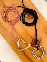 Collares de cordón de símil gamuza con dijes metálicos en forma de corazón y formas geométricas, adornados con pequeñas cuentas plateadas a lo largo del cordón.