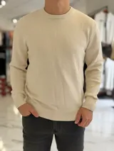 Buzo de hilo color beige claro, con cuello redondo y diseño clásico de manga larga.