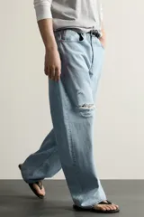 Jeans de corte barrel fit confeccionados en denim de algodón rígido, con diseño de cinco bolsillos y cierre frontal con botones. Presentan un lavado claro con efecto de roturas en las perneras.