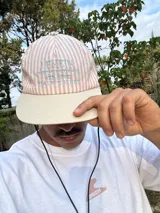 Gorro de visera curva con diseño a rayas verticales en tonos rosado pálido y blanco. La corona tiene un bordado en azul claro con texto. La visera es de color beige claro. Incluye un cordón negro ajustable para colgar del cuello.