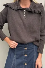 Sweater de punto color marrón, con cuello tipo babero con volados y cierre frontal parcial con tres botones. Presenta mangas largas con puños acanalados y diseño holgado.