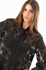 Blusa negra de tejido semitransparente con diseño floral en relieve, cuello alto con lazo frontal y mangas largas con volados en los puños.