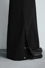 Pantalón de vestir gris oscuro, corte ancho (flare fit) y tiro alto, con bolsillos laterales y falsos bolsillos de vivo en la parte trasera. Presenta un cierre frontal con cremallera y gancho oculto, y botones a presión ocultos en el bajo.