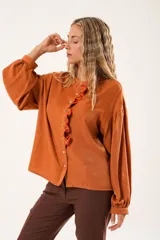 Camisa color naranja óxido con cuello mao, mangas largas abullonadas y cierre frontal con botones. Presenta un volante vertical con borde rojo en el frente.