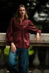 Blusa de satén color bordó con cuello redondo, cierre frontal con cordones y detalle de canesú con bordado calado y volados. Presenta mangas largas abullonadas con puños elásticos.