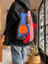 Campera de textura tipo corderito en color negro, con diseño de collage geométrico en la espalda que incluye parches en azul, naranja, marrón y franjas rojas y blancas. Posee capucha y mangas largas.