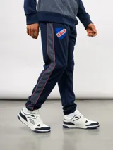 Pantalón deportivo de piqué color azul marino, con franjas laterales en contraste rojo y blanco. Presenta el escudo del Club Nacional de Football bordado en la pierna derecha y una etiqueta conmemorativa en la izquierda.