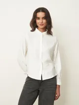Camisa de manga larga en color blanco, con cuello mao y detalle de volados sutiles en el borde. Presenta cierre frontal oculto con botones y puños con volados.