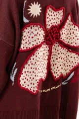 Saco de punto color vino con diseño holgado, cuello redondo y mangas largas. Presenta un gran bordado central en la espalda con forma de flor en tonos crema y rojo, acompañado de un pequeño bordado de sol y una frase bordada en la parte inferior.