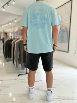 Remera celeste oversize con estampa en la espalda de un paisaje marino con palmeras y olas dentro de un círculo, acompañada de texto.