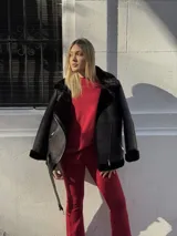 Campera tipo gamulán de cuero sintético negro con interior y detalles de corderito al tono. Presenta cierre frontal diagonal, cuello amplio, bolsillos laterales con cierre y cinturón ajustable en el ruedo.