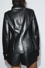 Chaqueta entallada de efecto piel en color negro, con cuello subido y cierre frontal cruzado con botones metálicos dorados. Cuenta con manga larga con trabillas en los puños y hombreras decorativas.