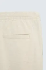 Pantalón de corte relajado color beige confeccionado en tejido de mezcla de algodón y lino. Posee cintura elástica ajustable con cordón, bolsillos frontales y bolsillos traseros de vivo.