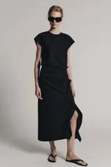 Vestido midi negro con cuello redondo y manga sisa, confeccionado por Zara Woman Collection. Presenta un diseño drapeado en la parte delantera y un bajo asimétrico con un voluminoso volante lateral. Incluye un pequeño cierre de botón en la espalda.