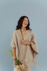 Ruana tejida a mano en lana merino color beige, con textura de punto y detalle de flecos en el ruedo. Presenta sutiles destellos brillantes integrados en el tejido.