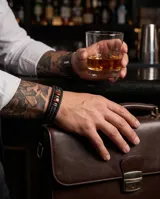 Pulsera de diseño masculino compuesta por una banda de cuero sintético trenzado en color negro, con detalles centrales de acero y piedras naturales ojo de tigre.