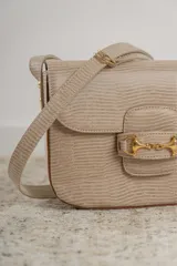 Cartera tipo bandolera de cuero con textura grabada, color beige. Presenta un detalle de herraje metálico dorado en la solapa frontal y placa con logo de la marca. Incluye correa ajustable para llevar al hombro o cruzada.
