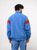 Buzo deportivo de nylon con medio cierre frontal y cuello alto. Presenta un diseño de bloques de color en azul, blanco y bordó, con el escudo del Club Nacional de Football y el logo de Umbro en el pecho.