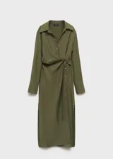 Vestido midi de diseño cruzado en color verde oliva, con cuello camisero, cierre frontal de botones y lazo lateral para ajustar en la cintura. Presenta mangas largas con puños abotonados y una abertura lateral pronunciada en el bajo.
