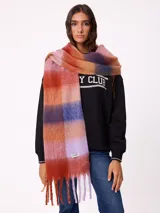 Bufanda de tejido suave con diseño de cuadros grandes en tonos naranja, rojo, violeta y lila. Presenta flecos en los extremos y una etiqueta pequeña con el logo de la marca Rusty.