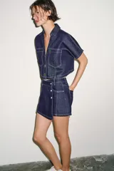 Camisa corta de denim de manga corta, con cuello solapa, escote en V y bolsillos delanteros de plastrón. Cuenta con costuras en contraste y cierre frontal con botones metálicos.