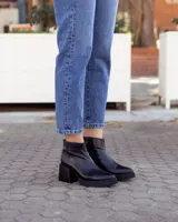 Botín de cuero negro, estilo botineta, con taco ancho y plataforma. Presenta un diseño liso y brillante, con cierre lateral.