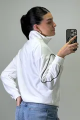 Buzo deportivo color beige con cuello alto y cierre frontal parcial. Presenta costuras decorativas con ribetes blancos en el pecho y a lo largo de las mangas. Posee puños y ruedo elásticos.