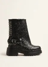 Bota corta de cuero negro con diseño de polaina superpuesta, detalle de tachas metálicas en el lateral y correa con hebilla ajustable. Cuenta con suela track de goma y taco cuadrado.