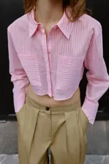 Camisa corta rosa con rayas verticales finas, cuello solapa, manga larga y bolsillos delanteros de plastrón. Cierre frontal con botones ocultos.