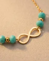 Tobillera de cadena fina de acero con baño de oro, con dije central de símbolo de infinito y cuentas de piedra turquesa.