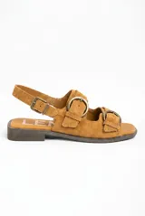 Sandalias planas de gamuza color tostado, con múltiples tiras decoradas con grandes hebillas metálicas en tono bronce viejo y tira ajustable en el talón.