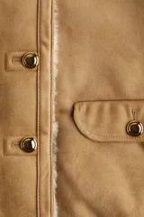 Chaqueta corta de gamuza sintética color tostado, con forro interior de peluche color crema. Presenta cuello redondo, cierre frontal con botones metálicos y dos bolsillos delanteros con solapa y botón.