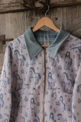 Campera de lona estampada con diseño de caballos y flores, con cuello de pana en color gris azulado y cierre frontal metálico. El interior está forrado en tela de viyela a cuadros.