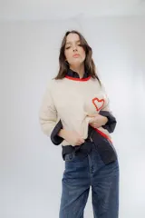 Buzo de punto color crema con detalles en rojo en el cuello, puños y ruedo. Presenta un bolsillo frontal en el pecho con un corazón bordado en relieve.