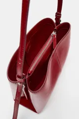 Bolso de hombro en formato mini color rojo, con detalle de pespuntes centrales y asa ajustable. Cuenta con compartimento interior con cremallera y doble compartimento exterior con cierre imantado.