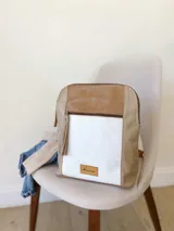 Mochila de diseño tricolor con paneles en beige, blanco con textura tipo reptil y detalles en cuero sintético marrón. Cuenta con un bolsillo frontal con cierre, correas ajustables y etiqueta de marca en la parte inferior.