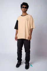 Remera de corte oversize en color beige, con cuello redondo en contraste negro y una manga en color negro. Presenta un pequeño estampado circular en el pecho con la frase 'Don't worry, be happy'.