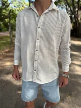 Camisa de lino blanca con rayas verticales beige, de manga larga, cuello camisero y cierre frontal de botones.