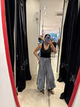 Pantalón de corte amplio y fluido, color gris con acabado satinado, con cintura elástica y lazo ajustable. Presenta un diseño con paneles laterales que aportan volumen y movimiento.