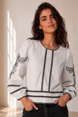 Blusa blanca con detalles de bordados florales grises en las mangas y pasamanería oscura en el frente y puños.