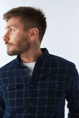 Camisa de pana a cuadros en tonos azul y bordó, de corte regular, manga larga, con cartera de botones y bolsillo frontal.