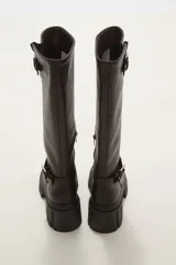 Bota de caña alta color negro, confeccionada en eco cuero, con suela track de goma y detalle de hebilla metálica en el tobillo y en la parte superior de la caña.