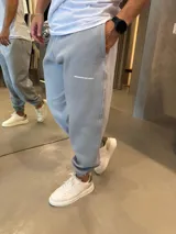 Pantalón de jogging gris claro con puños elásticos en los tobillos, bolsillos laterales y logo de la marca estampado en la pierna. Confeccionado en algodón premium con textura suave.
